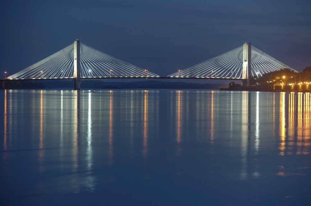 A cable-stayed bridge lit up at dusk over the Mississippi River, its lights reflected in the water.